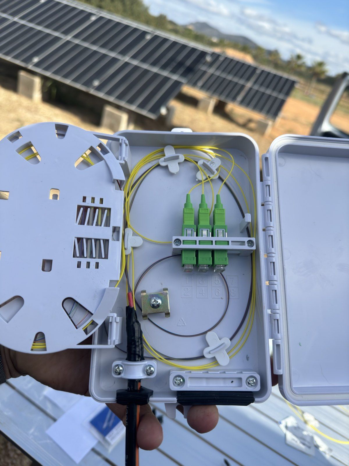 Geöffnete Glasfaser-Verteilerbox mit grünen SC/APC-Steckern vor Solarmodulen auf einer Finca