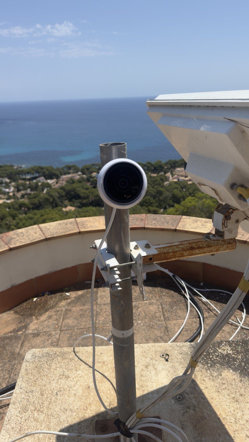 UniFi-Kamera an einem Mast auf einer Dachterrasse mit Blick aufs Meer, daneben eine Richtfunkantenne