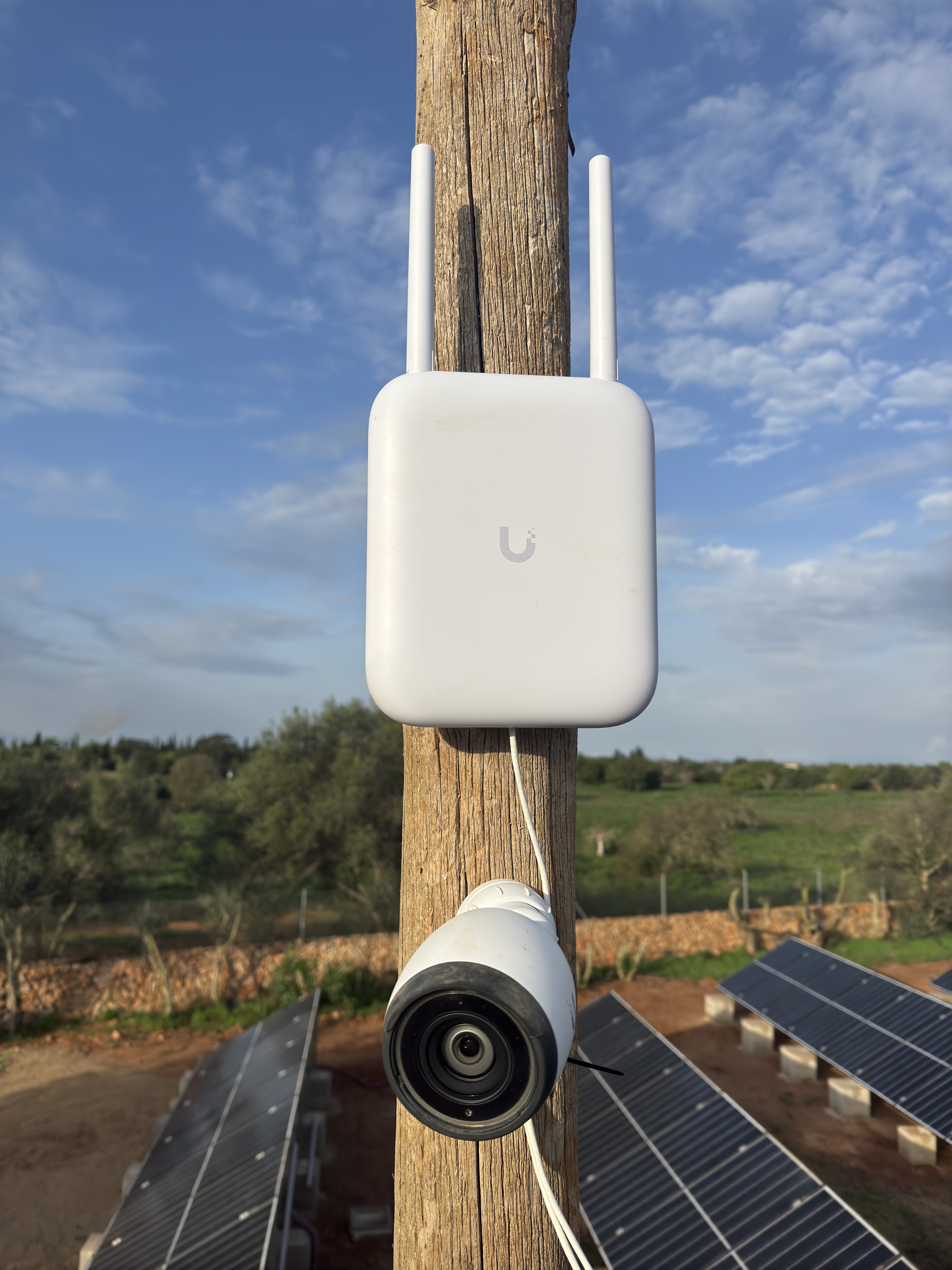 JumpIT UNIFI Ein weißes drahtloses UNIFI-Mallorca-Gerät mit zwei Antennen und eine Überwachungskamera sind an einem Holzpfahl montiert. Darunter sind Sonnenkollektoren zu sehen. Der Hintergrund zeigt grüne Felder und einen teilweise bewölkten Himmel.